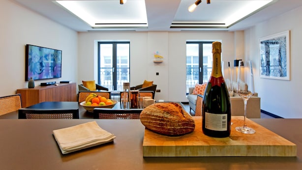 A modern living room with a dining area, featuring a wooden table with a bottle of champagne and a glass, a loaf of bread, and a fruit bowl. - 2-bed Superior Apt 415 (London)