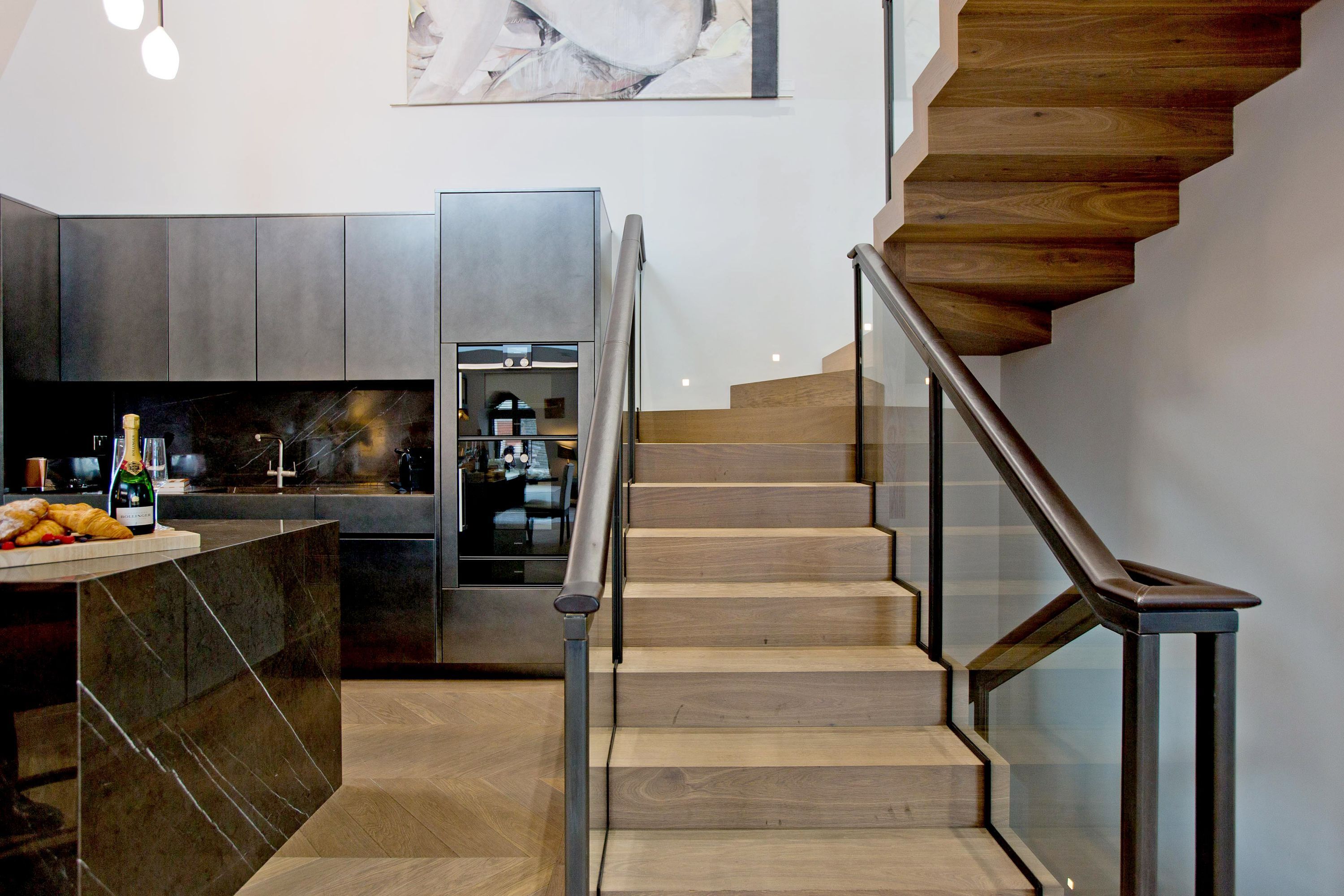 A modern kitchen with a central island, a built-in refrigerator, and a wooden staircase with glass railings.