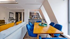 A modern dining area with a wooden table and blue chairs, a light blue sofa with yellow cushions, and a kitchen in the background. - 2-bed Superior Apt 416 (London)
