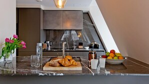 A modern kitchen with a marble countertop, a wooden cutting board with croissants, a bowl of fruit, and a vase of flowers. - 2-bed Superior Apt 416 (London)