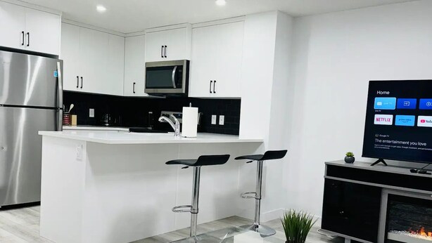 Eine moderne Küche mit weißen Schränken, einem Edelstahlkühlschrank, einer Mikrowelle und einem Flachbildfernseher. Eine Kochinsel mit Glasplatte, zwei Barhocker und ein kleiner Topfpflanze sind ebenfalls vorhanden.