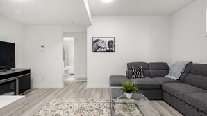 Ein modernes Wohnzimmer mit einem Ecksofa, einem Glastisch mit Pflanze, einem Flachbildfernseher und einem gemusterten Teppich.