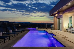 Piscina al aire libre con vistas a montañas al atardecer, rodeada de sillas de descanso y un edificio moderno.