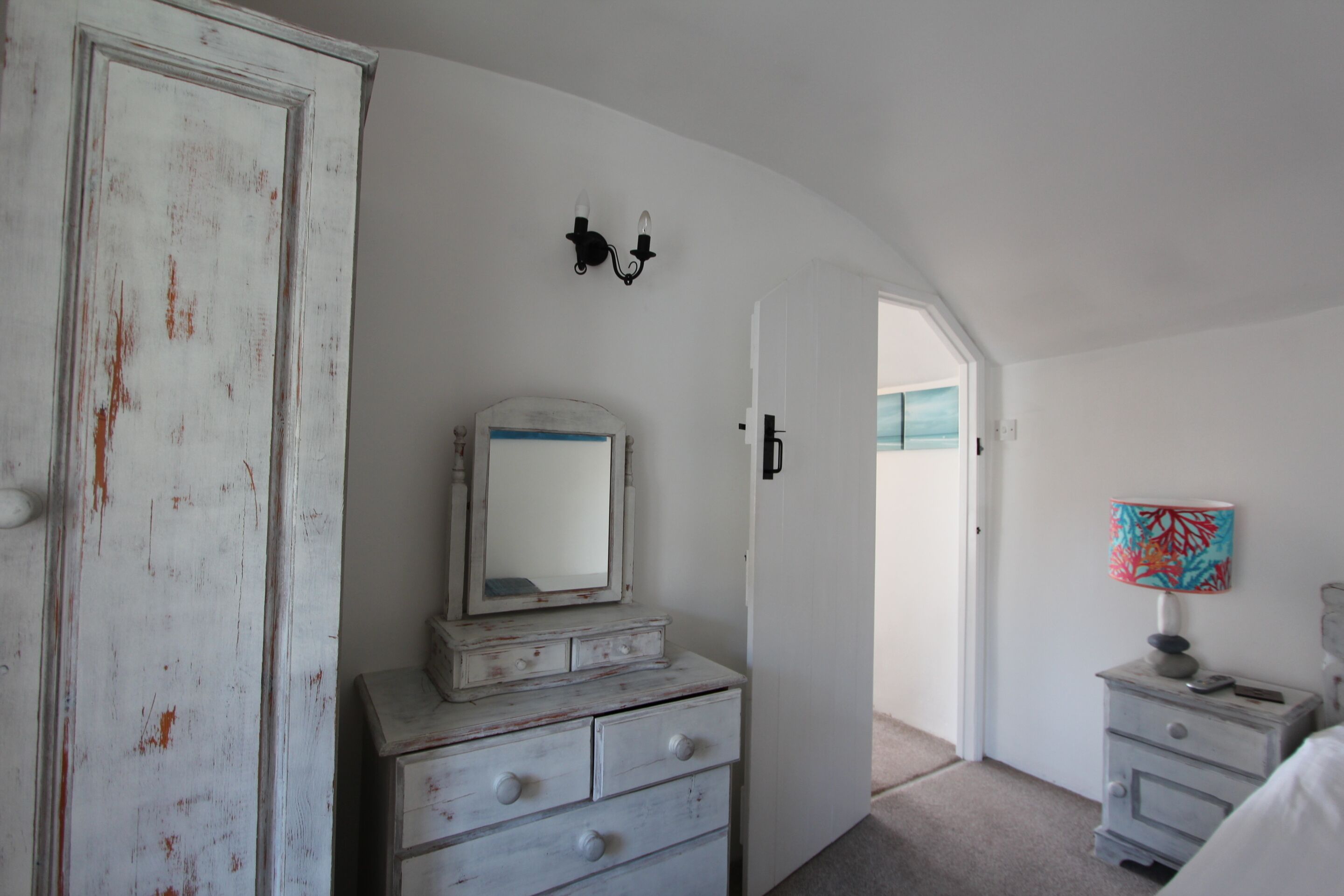 A bedroom with a white dresser, a mirror, a lamp, and a door leading to another room.