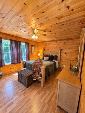 A wooden cabin interior with a bed, a desk, a chair, and a window with curtains.