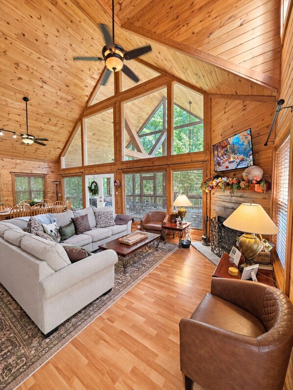 A spacious living area with a high ceiling, large windows, and a fireplace.