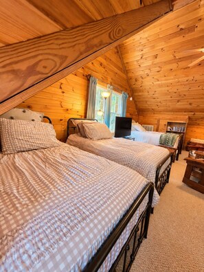 A cozy attic bedroom with two beds, a desk, and a window with curtains.