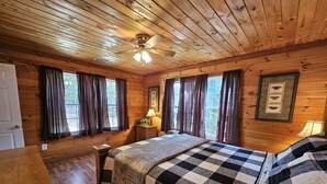 Camera da letto in una baita di legno con un letto, comodini, una ventola a soffitto e una finestra con tende.