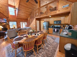 A wooden cabin interior with a dining area, kitchen, and a staircase leading to an upper level.
