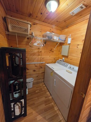 A compact laundry room with wooden walls, a washing machine, a drying rack, and a small trash bin.