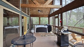 Un'area esterna riparata con travi in legno, ventilatori a soffitto e ampie finestre che offrono vista sugli alberi. Si notano tavoli e sedie in metallo, un barbecue e una vasca idromassaggio.