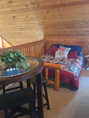 A cozy room with a wooden ceiling, a round glass table with chairs, a bed with a quilt, and a potted plant.