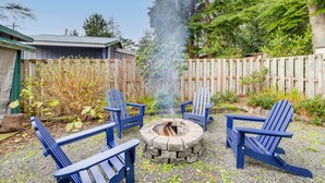 A fire pit with four Adirondack chairs around it, set on a gravel surface with a wooden fence and greenery in the background. - Rockaway Beach Home, Walk to Ocean, Pet Friendly! (Rockaway Beach)