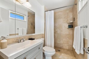 Une salle de bain avec un lavabo blanc, un miroir et des toilettes. On y trouve un rideau de douche et un porte-serviettes.