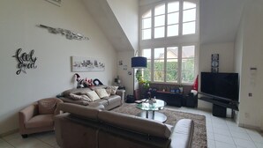 A spacious living room with a high ceiling, a large window, a sofa, a coffee table, and a flat-screen TV.
