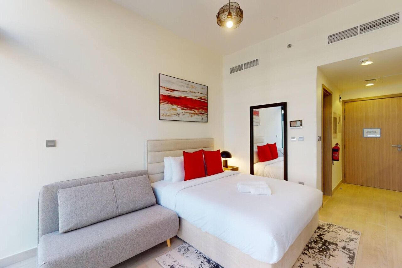 A modern hotel room with a large bed, a grey sofa, a red and white abstract painting, a mirror, and a wooden door.