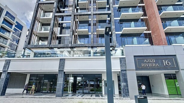Un edificio moderno con fachada de vidrio, un letrero que dice "AZIZI RIVIERA 16" y una entrada pavimentada donde se ve un extintor.