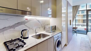 Una cocina moderna con estufa a gas, pileta y microondas, lavadora y vista a un edificio a través de amplios ventanales.
