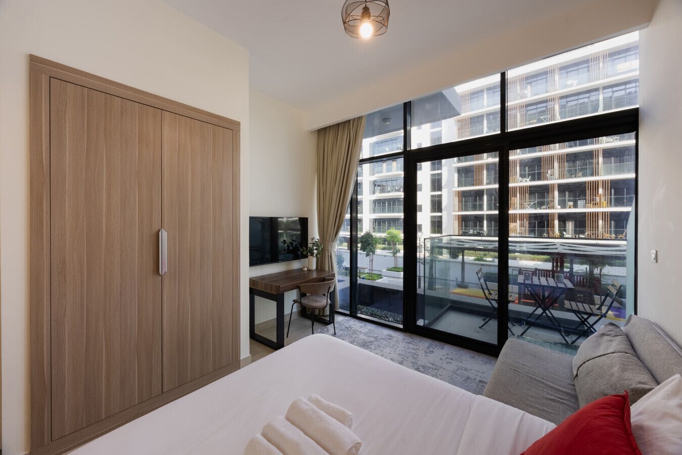 A hotel room with a large bed, a desk with a TV, a balcony with a view of a modern building, and a wardrobe.