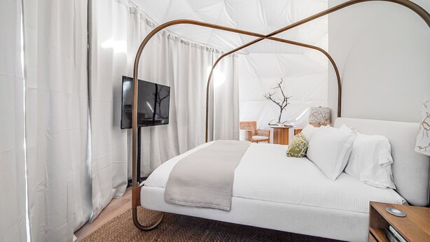 A modern bedroom with a four-poster bed, a flat-screen TV mounted on the wall, and a small side table with a lamp and decorative items.