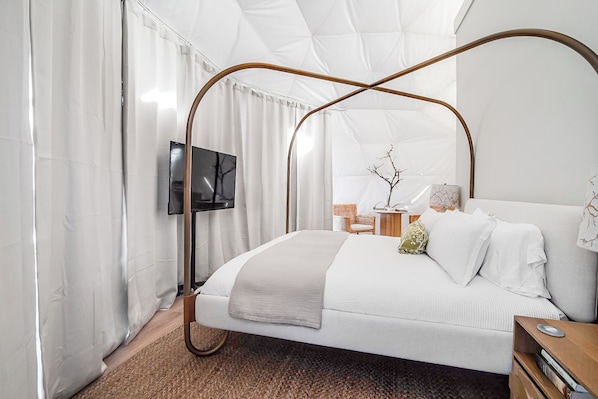 A modern bedroom with a four-poster bed, a flat-screen TV mounted on the wall, and a small side table with a lamp and decorative items.