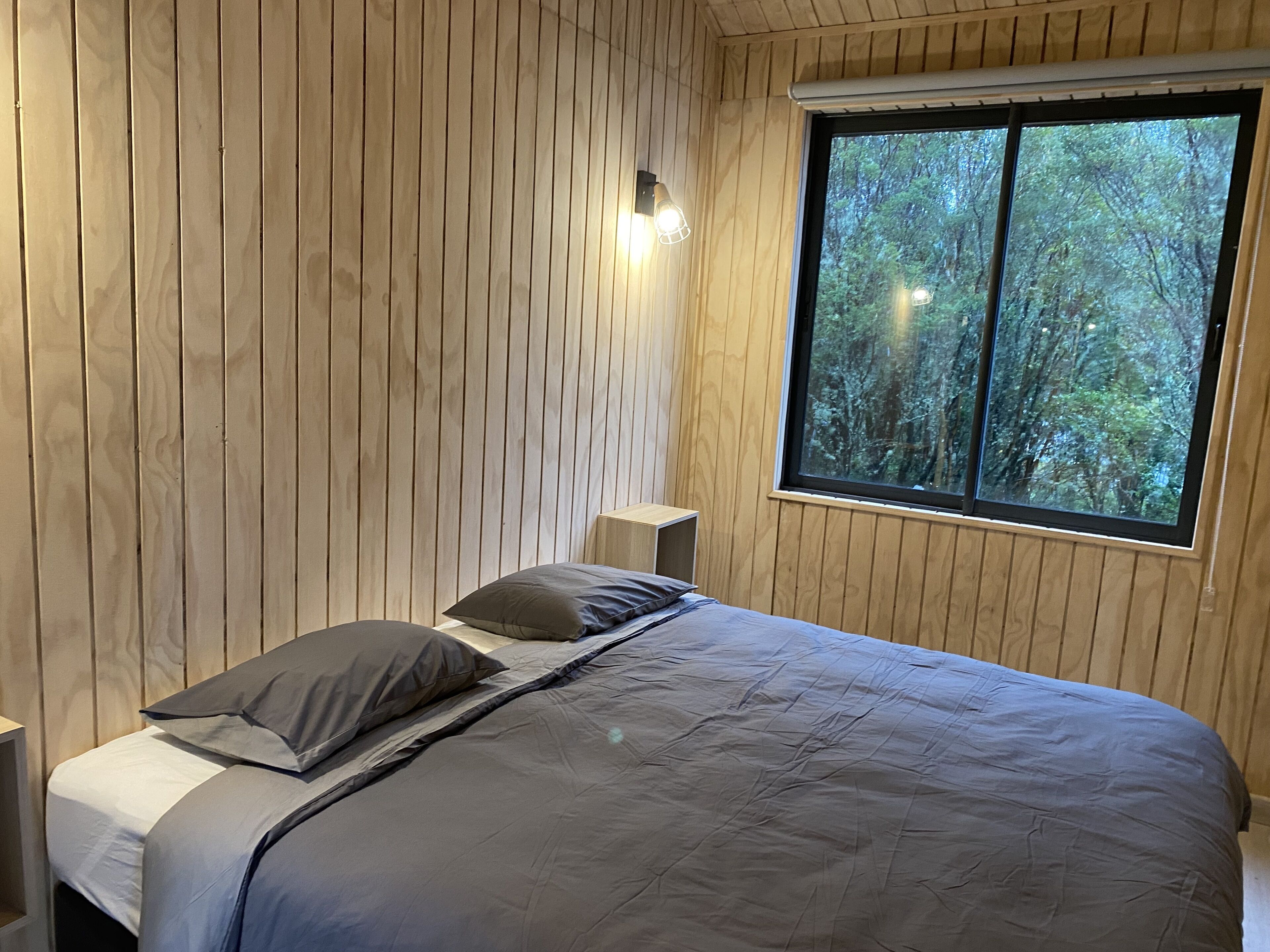 Un dormitorio con paneles de madera, una cama con ropa de cama gris, una mesita de noche y una ventana con vista a árboles.