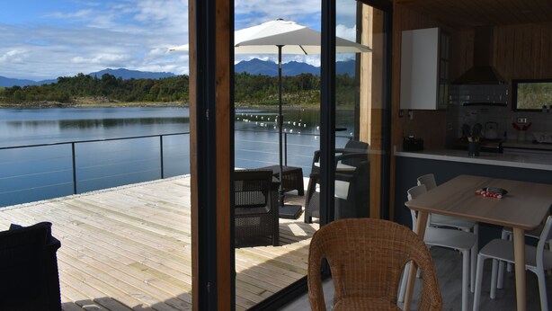 Eine moderne Küche mit einem großen Fenster, einem Essbereich mit Tisch und Stühlen sowie Blick auf einen See mit Bergen im Hintergrund.