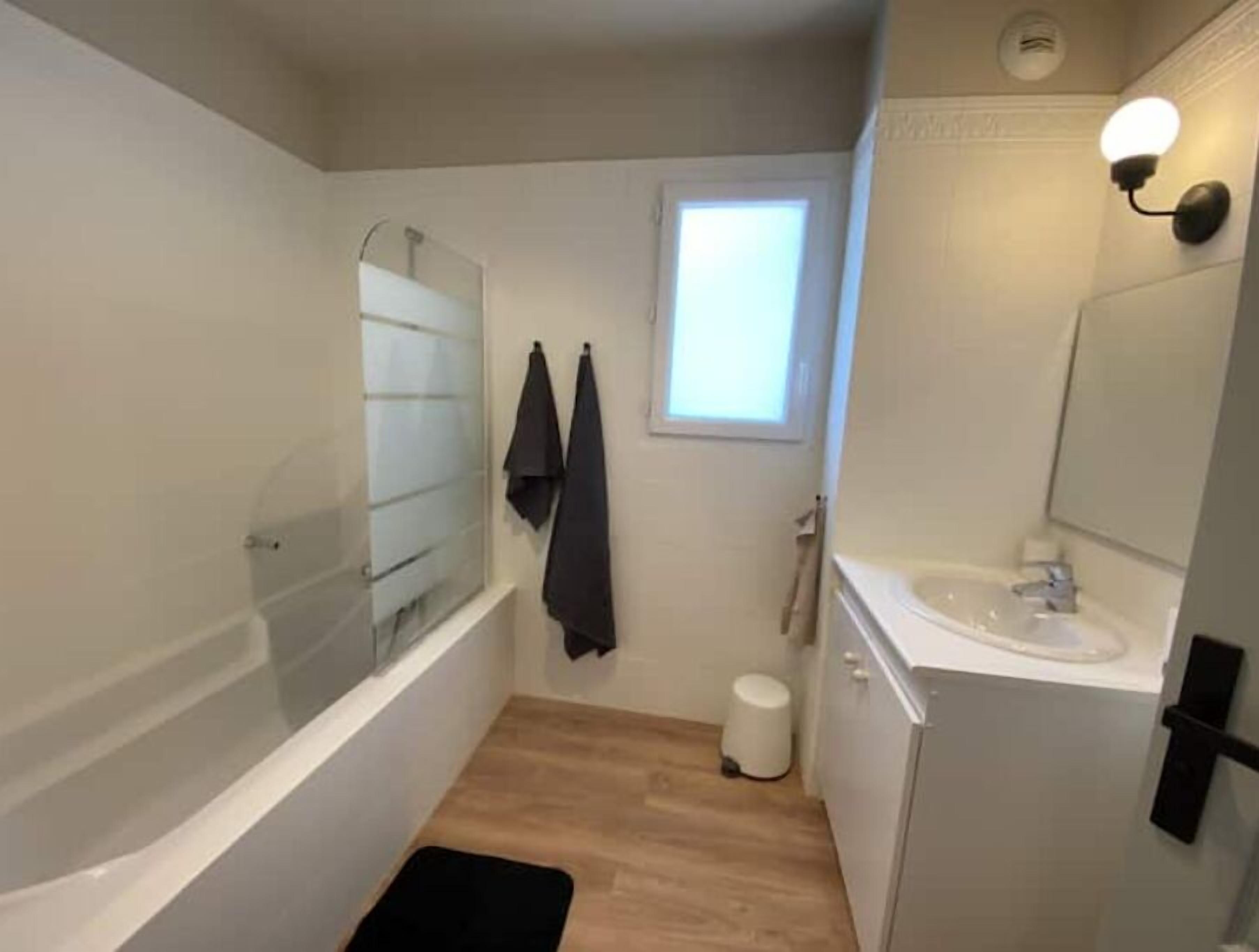 A small, compact bathroom with a bathtub, a sink, and a mirror. There is a window providing natural light and a towel hanging on the wall.