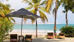 A beachside seating area with wooden lounge chairs and a large umbrella, overlooking a beach with palm trees and wind turbines in the distance. - Pousada Les Alizes (Amontada)