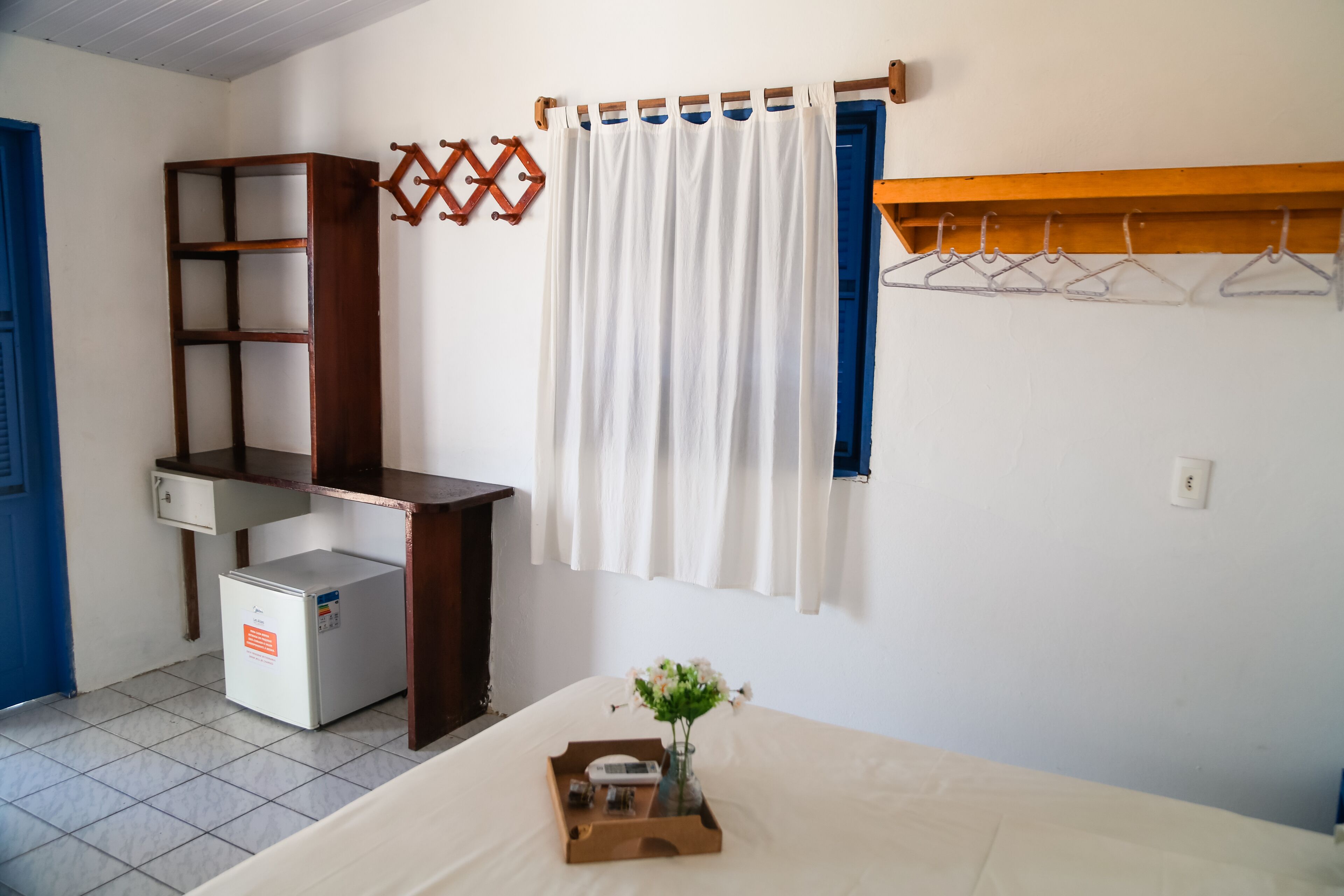Een slaapkamer met een bed, een kleine witte koelkast, een houten bureau, een wit gordijn en een houten kapstok.