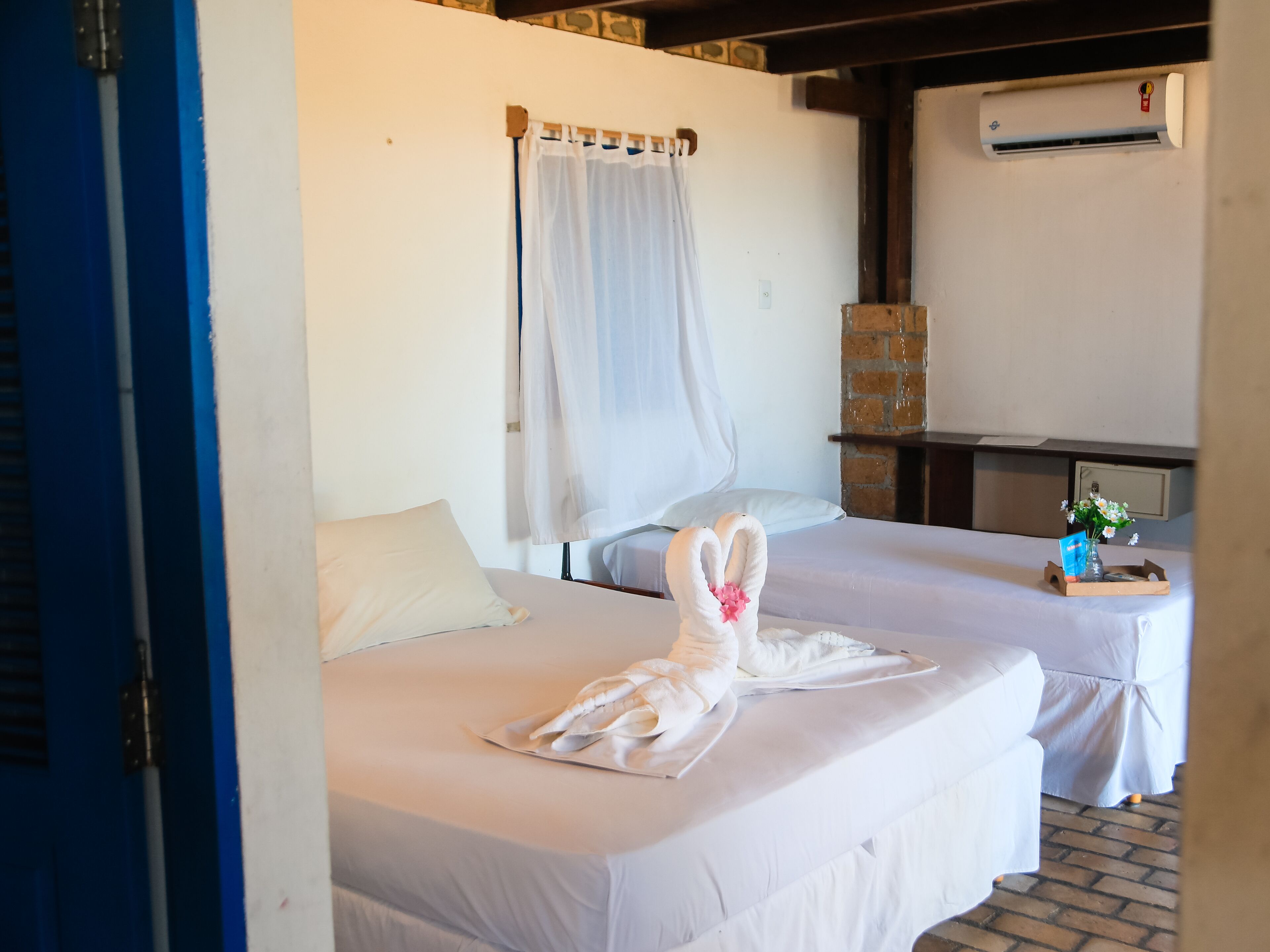 A hotel room with two beds, a white curtain, an air conditioning unit, and a small table with a vase of flowers.