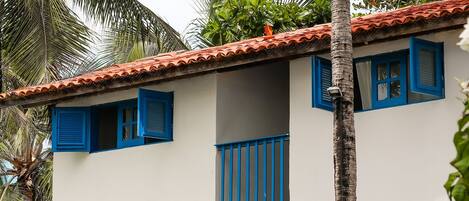 A two-story building with white walls and blue shutters, surrounded by lush greenery and palm trees.