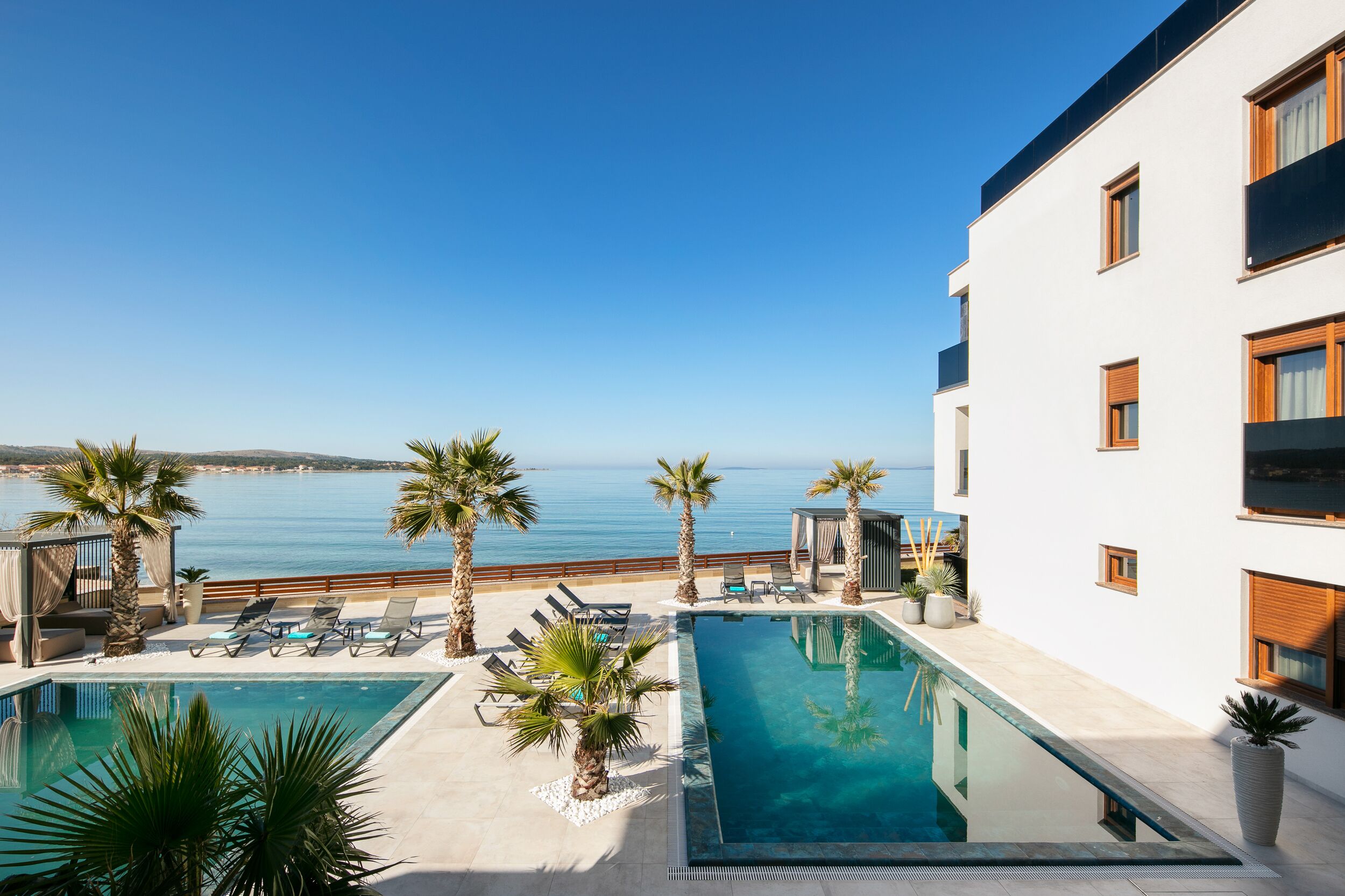 Zona de piscina con dos piscinas, palmeras, sillas de descanso y vistas al mar.