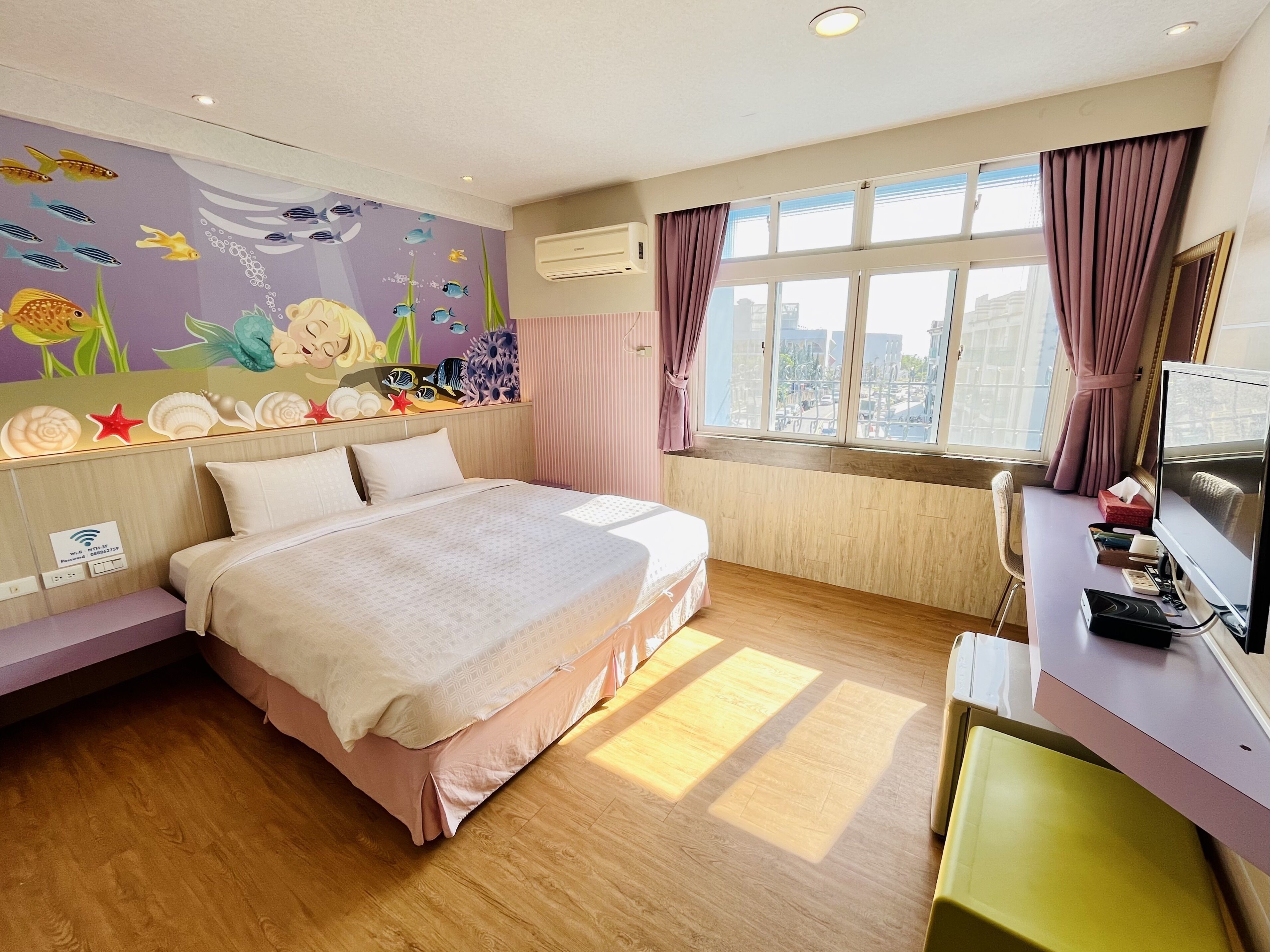 Een hotelkamer met een bed, een bureau met een computer, een raam met gordijnen en een muurschildering.