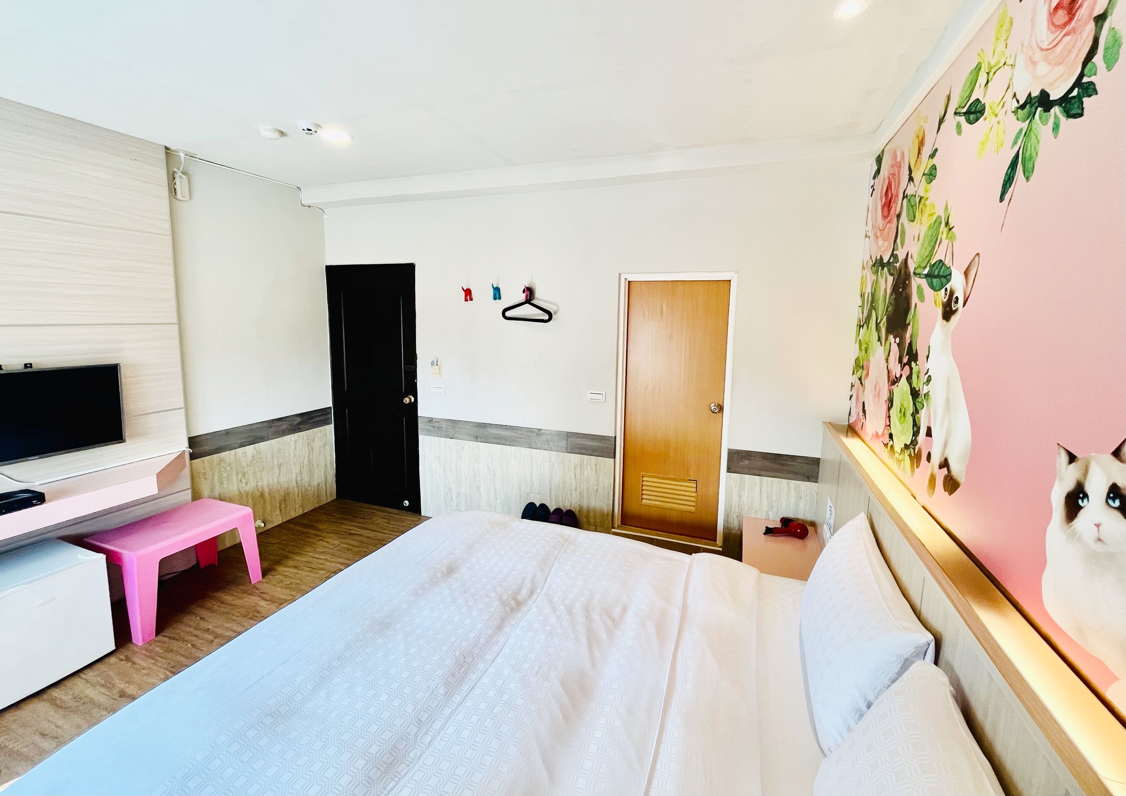 Een hotelkamer met een bed, een televisie, een roze krukje, een houten deur en een muur met een bloemschilderij.
