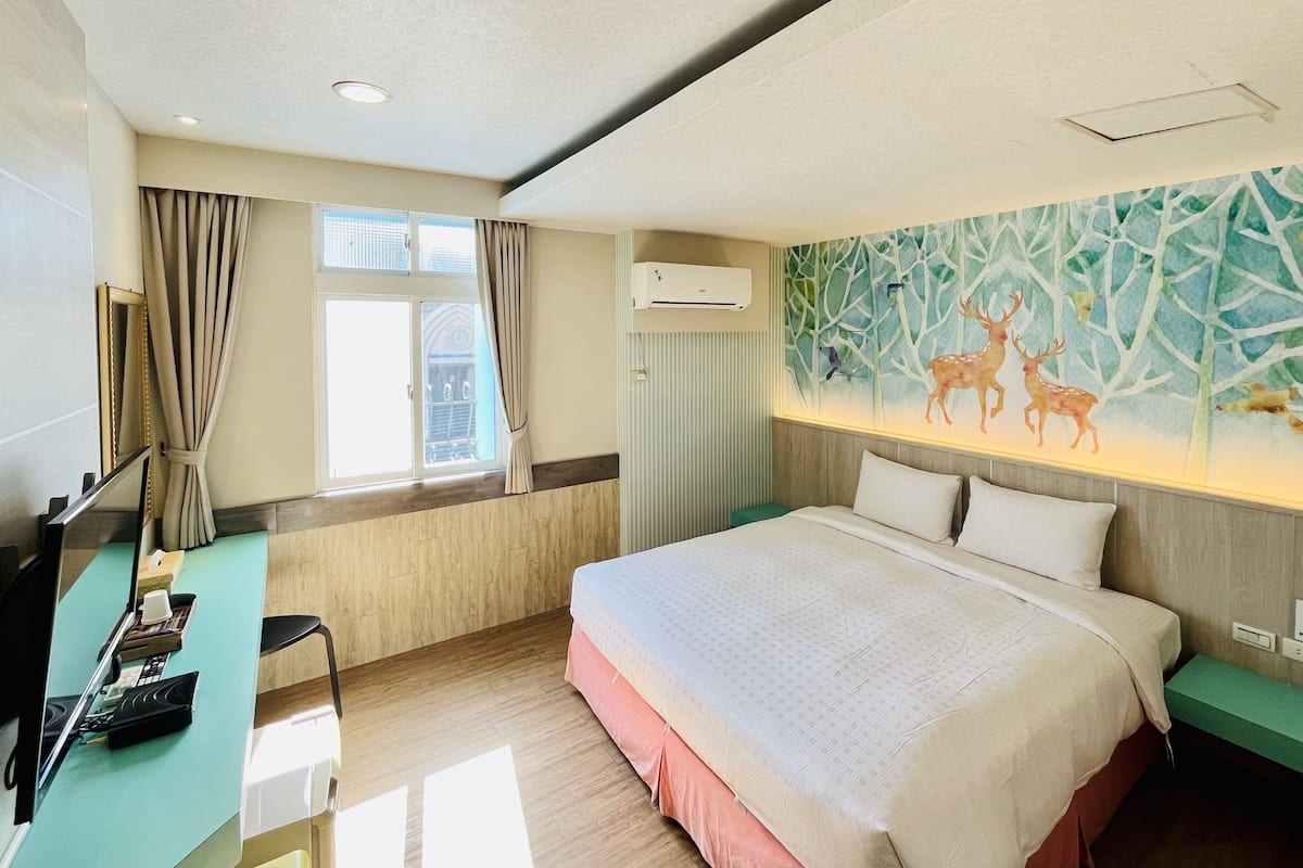 A hotel room with a large bed, a desk with a computer, a window with curtains, and a wall mural of deer.