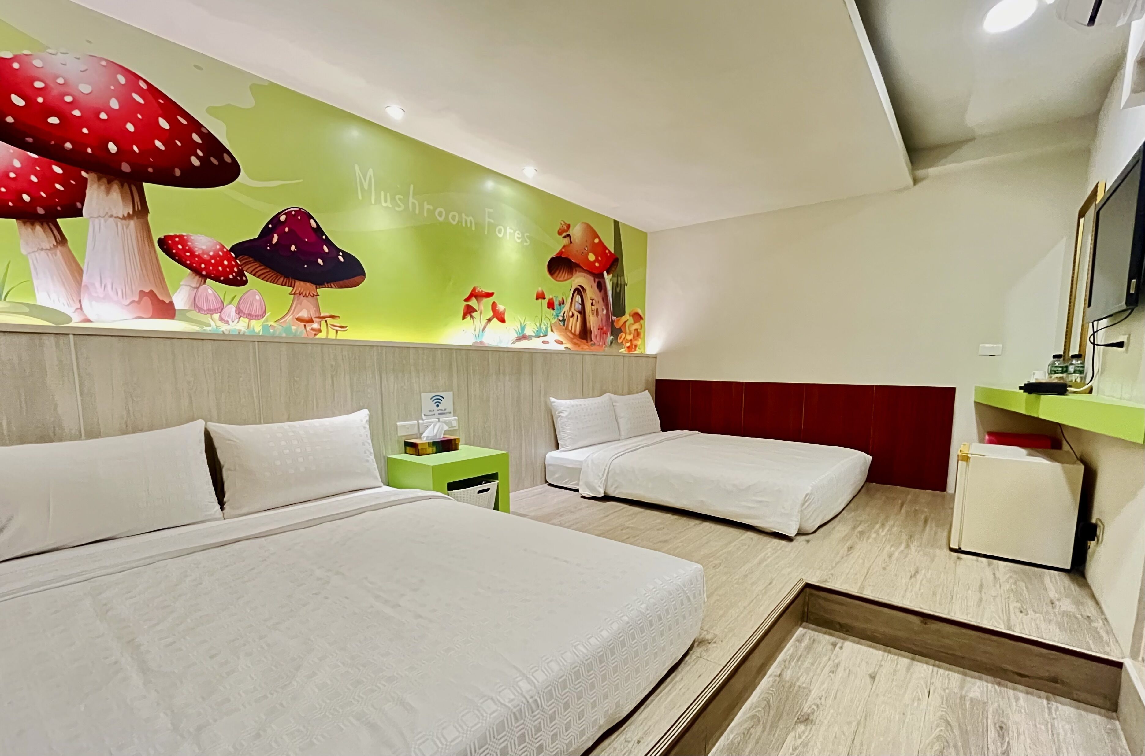A hotel room with two beds, a green wall mural, a small nightstand, and a red accent wall.