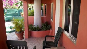 Área de estar externa coberta com mesa e cadeiras, cercada por plantas em vasos e vista para um espaço verde com escadas.