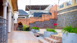 A courtyard with a tiled floor, a brick wall, and a stone wall. There are steps leading up to a raised platform with potted plants. - Inviting 6-bedrooms Apartment in Kigali (Kigali)