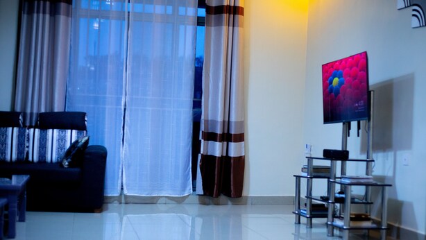 A living room with a sofa, a TV on a stand, and sheer curtains. - Inviting 6-bedrooms Apartment in Kigali (Kigali)