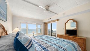 Una camera da letto con un letto grande, una televisione su un mobile in legno, una ventola a soffitto e vista sull'oceano dalle finestre.