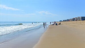 Une plage où l’on voit des gens se promener et jouer, un rivage sablonneux et des bâtiments en arrière-plan.