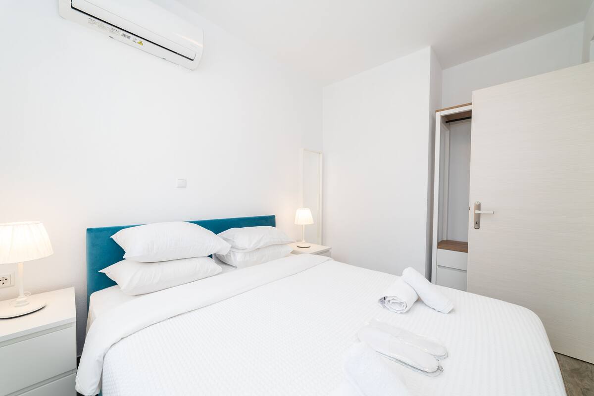 A hotel room with a bed, white linens, a blue headboard, a bedside lamp, a wall-mounted air conditioner, and a door.