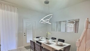 A dining area with a white table set for four, surrounded by chairs. There are decorative lights above the table and a staircase to the right.