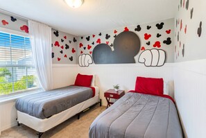 A room with two beds, a window with curtains, a radiator, and Mickey Mouse-themed wall decorations.