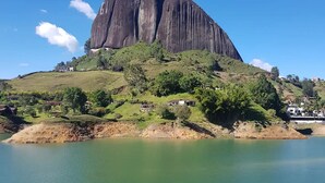 A large, dark rock formation with a small building on top, surrounded by greenery and a body of water. - Unique Lake House breathtaking views (Guatape)