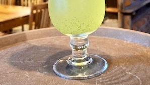 Un cóctel en un vaso alto con espuma, sobre una mesa en un restaurante con mobiliario de madera y un ambiente cálido.