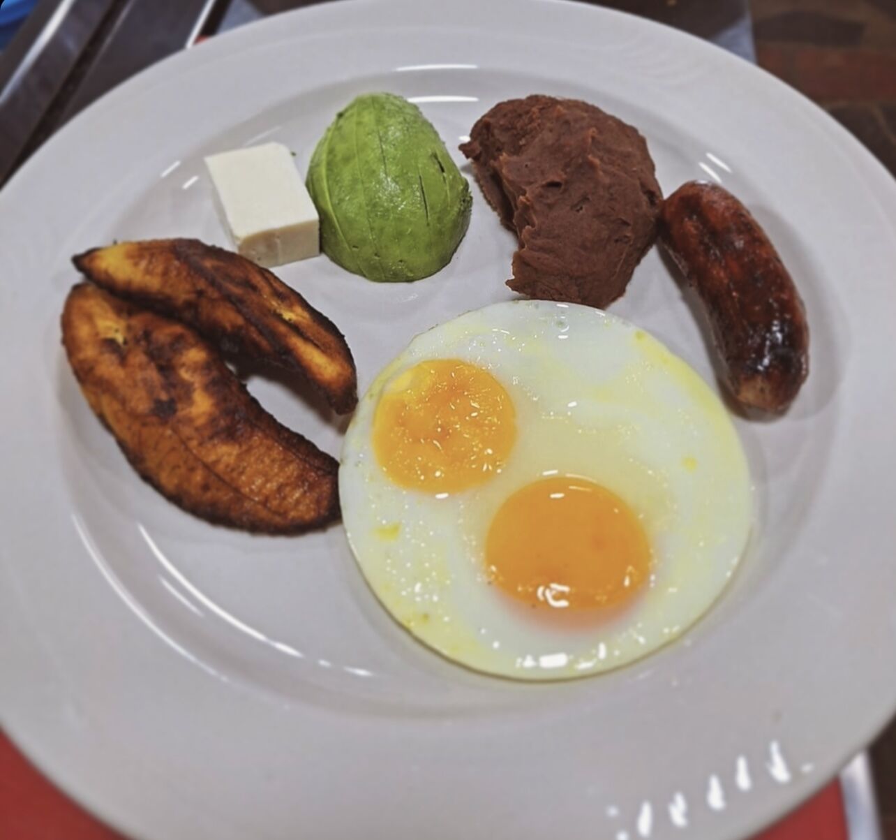 A plate with fried eggs, sausages, a piece of avocado, a slice of cheese, and grilled plantains.