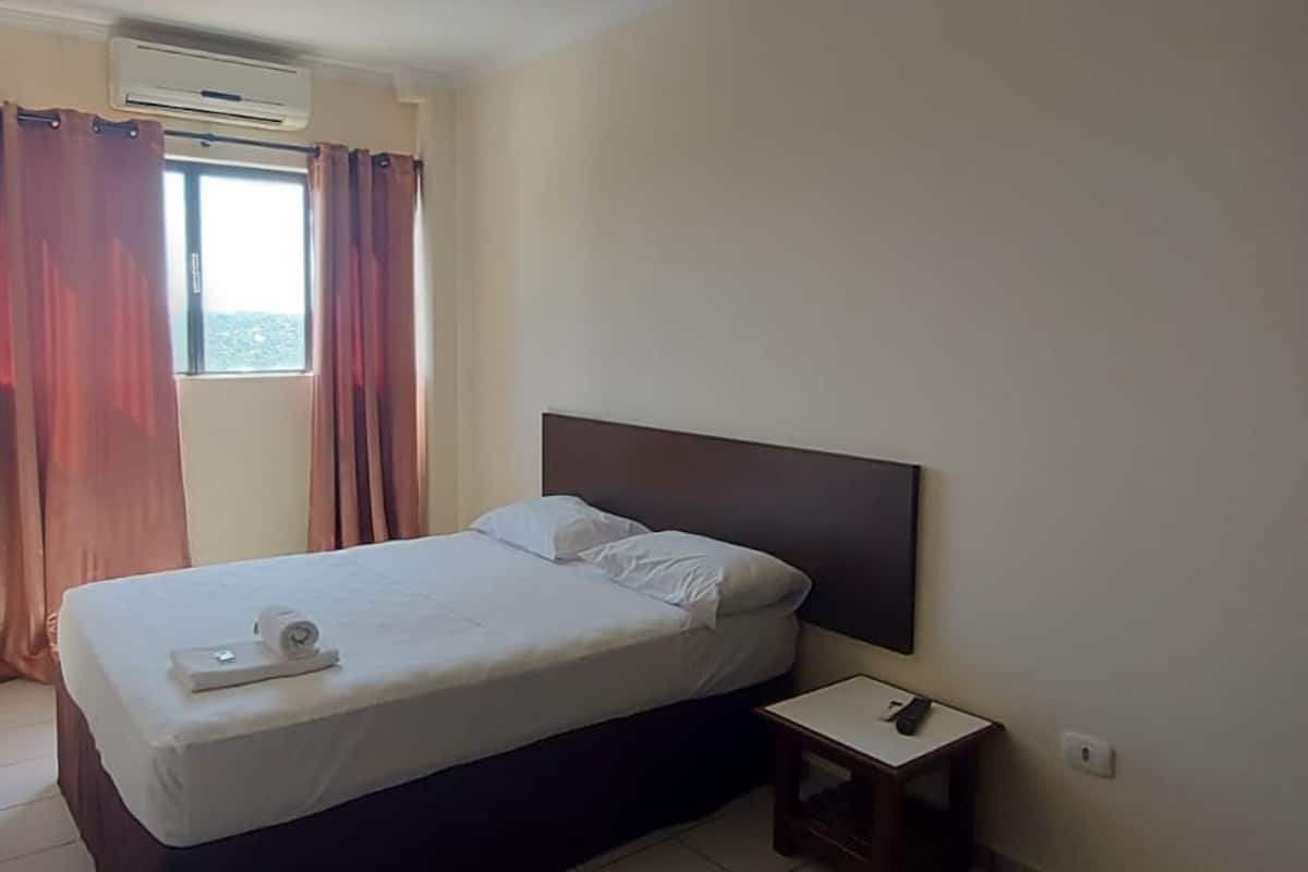 A hotel room with a bed, a nightstand, a window with curtains, and an air conditioning unit.