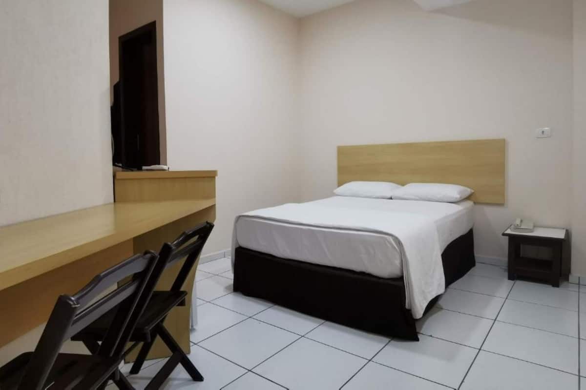 A hotel room with a bed, a desk, a chair, a TV, and a bedside table.